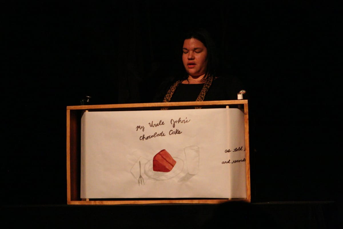 Ashley Minner standing at a podium, performing My Uncle John's Chocolate Cake. Photographed by Bill Haas.
